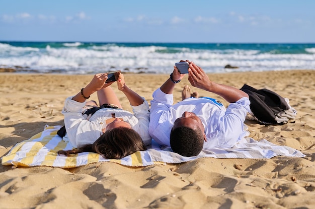 Joven pareja multicultural tirada en la playa usando teléfonos inteligentes