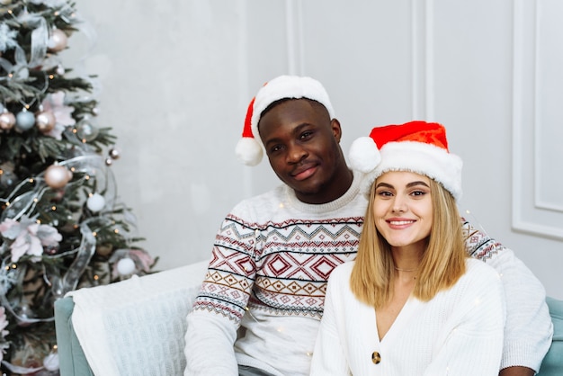 Una joven pareja multiétnica con gorros de santa está sentada en un sofá en un interior luminoso junto a un árbol. Hombre y mujer mirando a la cámara, sonriendo.