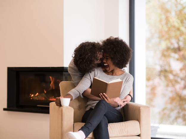 Joven pareja multiétnica hermosa abrazándose frente a la chimenea en casa cuando lee un libro en el día de otoño