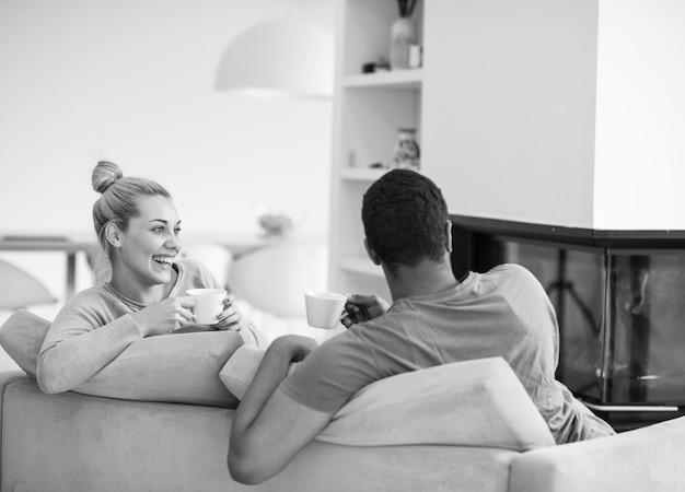 Joven pareja multiétnica romántica sentada en un sofá frente a la chimenea en casa, mirándose, hablando y bebiendo café