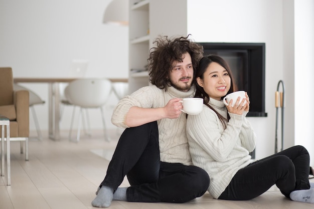 Foto joven pareja multiétnica romántica sentada en el suelo frente a la chimenea en casa, hablando y bebiendo café en el frío día de invierno