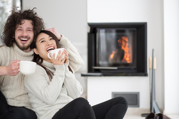 Joven pareja multiétnica romántica sentada en el suelo frente a la chimenea en casa, hablando y bebiendo café en el frío día de invierno