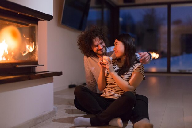 Joven pareja multiétnica romántica sentada en el suelo frente a la chimenea en casa, hablando y bebiendo té en la fría noche de invierno