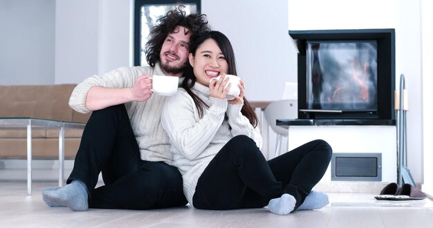 Joven pareja multiétnica romántica sentada en el suelo frente a la chimenea en casa, mirándose, hablando y tomando café en el día de otoño