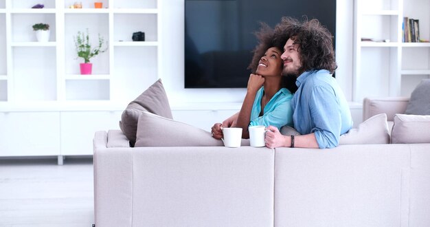 joven pareja multiétnica sentada en un sofá en casa bebiendo café, hablando, sonriendo.