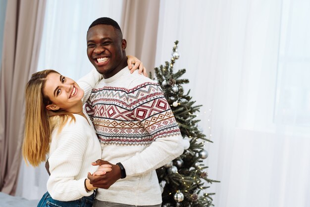 Joven pareja multinacional está bailando, riendo junto al árbol de Navidad. Encantadora pareja bailando en la sala de estar, celebrando la Nochebuena.