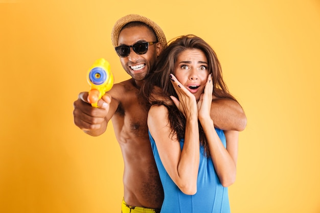 Foto joven pareja multirracial feliz divirtiéndose jugando con pistolas de agua aisladas en la pared naranja