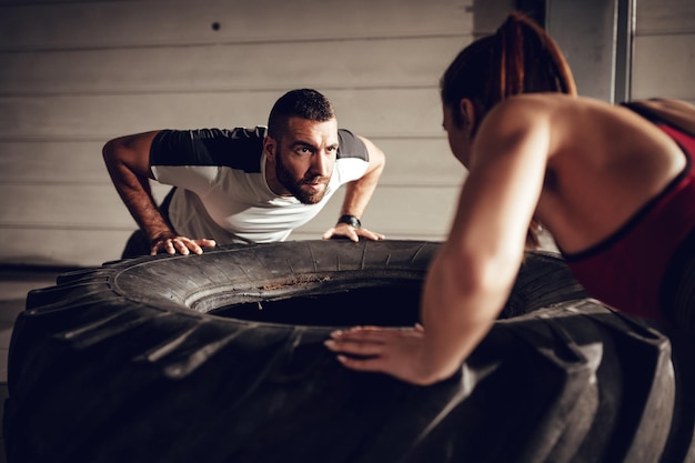 Joven pareja musculosa haciendo ejercicio push-up en un neumático en entrenamiento duro en el garaje.