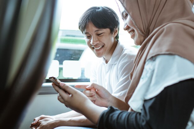 Una joven pareja musulmana sonriente mirando la pantalla del teléfono móvil juntos mientras está sentado en el autobús