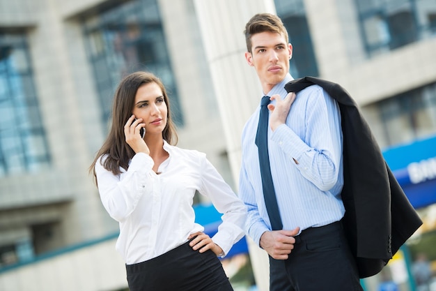 Joven pareja de negocios parada frente a edificios de oficinas y mujer de negocios hablando por teléfono móvil.