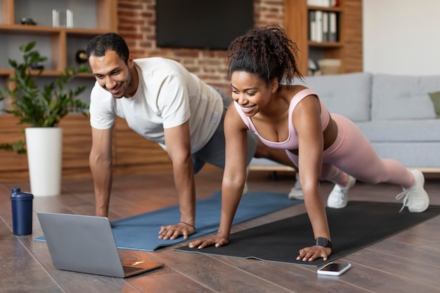 Una joven pareja negra sonriente con ropa deportiva ve un video en una laptop haciendo flexiones en el suelo juntos