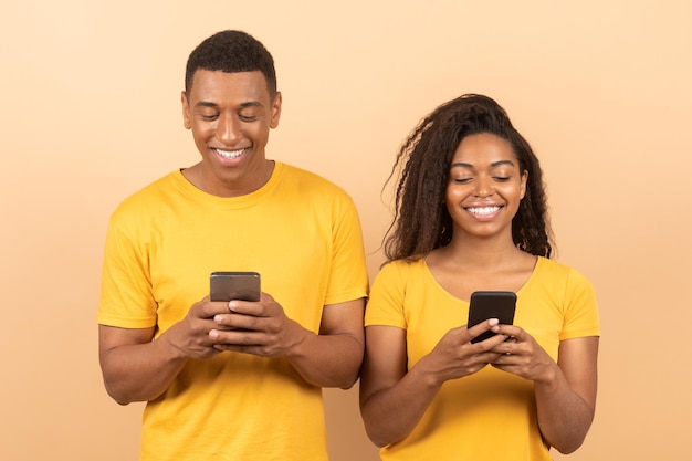 Joven pareja negra sosteniendo sus teléfonos inteligentes y jugando juegos o chateando en línea mirando aparatos y sonriendo