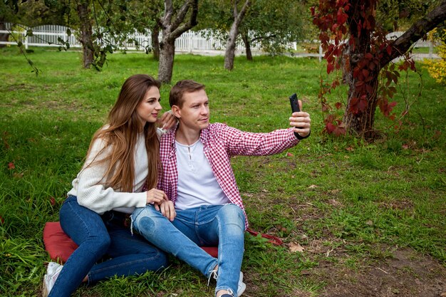 Una joven pareja pasa tiempo juntos en el parque, sentados en el césped, haciendo selfies en el teléfono. El chico con una chica con gadgets en el parque.