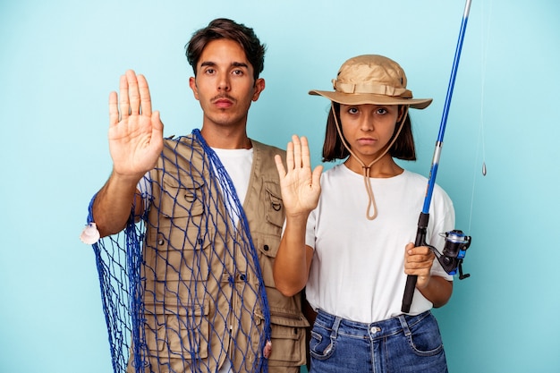 Joven pareja de pescadores de raza mixta aislado sobre fondo azul de pie con la mano extendida mostrando la señal de stop, impidiéndole.