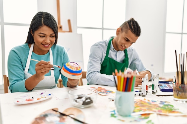 Joven pareja de pintores latinos sonriendo feliz pintando sentados en la mesa en el estudio de arte