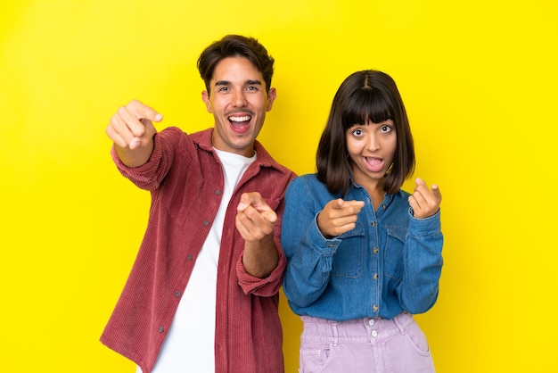 Una joven pareja de raza mixta aislada de fondo amarillo te señala con el dedo mientras sonríe