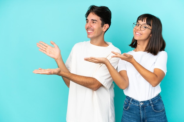 Joven pareja de raza mixta aislada de fondo azul extendiendo las manos a un lado para invitar a venir