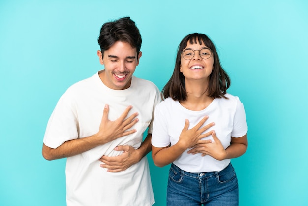 Joven pareja de raza mixta aislada de fondo azul sonriendo mucho mientras se pone las manos en el pecho