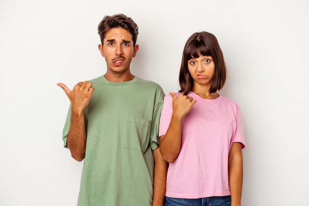 Una joven pareja de raza mixta aislada de fondo blanco sorprendida señalando con el dedo índice a un espacio de copia.