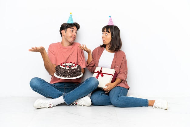 Joven pareja de raza mixta celebrando un cumpleaños sentada en el suelo aislada de fondo blanco haciendo un gesto sin importancia mientras levanta los hombros