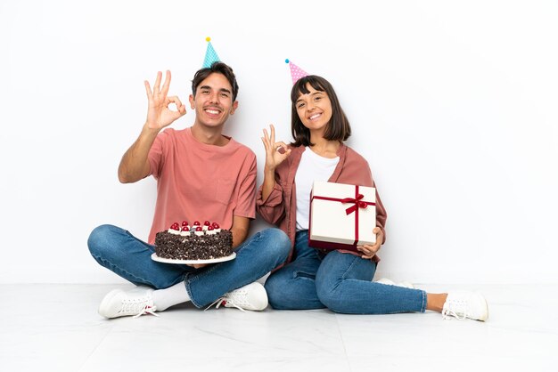 Una joven pareja de raza mixta celebrando un cumpleaños sentada en el suelo aislada de fondo blanco mostrando un signo de ok con los dedos