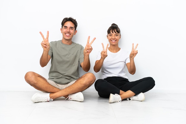 Una joven pareja de raza mixta sentada en el suelo aislada de fondo blanco sonriendo y mostrando el signo de la victoria con ambas manos
