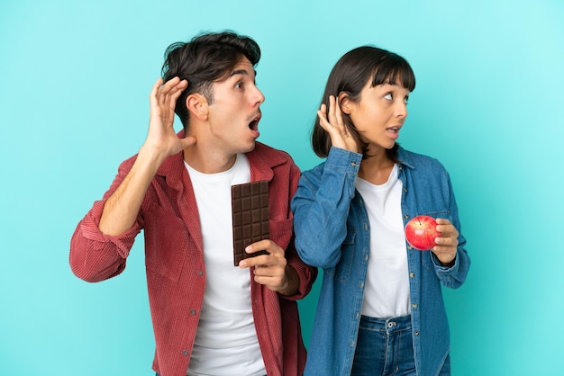 Joven pareja de raza mixta sosteniendo manzana y chocolate aislado en fondo azul escuchando algo poniendo la mano en la oreja