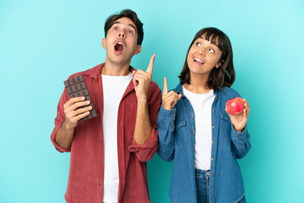 Joven pareja de raza mixta sosteniendo manzana y chocolate aislado en fondo azul pensando en una idea apuntando con el dedo hacia arriba