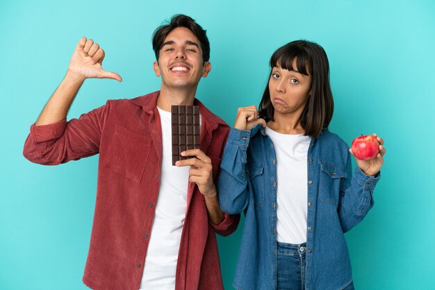 Joven pareja de raza mixta sosteniendo manzana y chocolate aislado sobre fondo azul orgulloso y satisfecho de sí mismo en el concepto de amarte a ti mismo