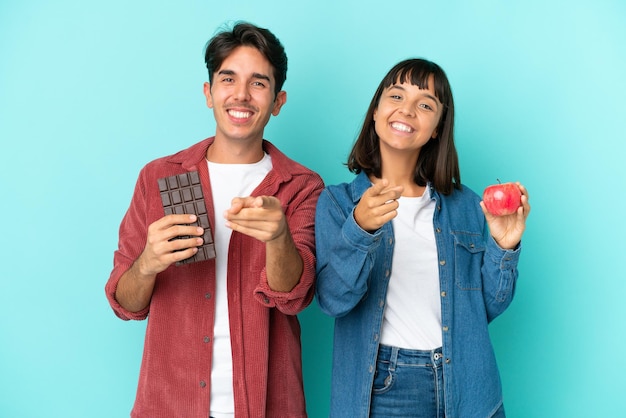 Joven pareja de raza mixta sosteniendo manzana y chocolate aislado sobre fondo azul te señala con una expresión de confianza