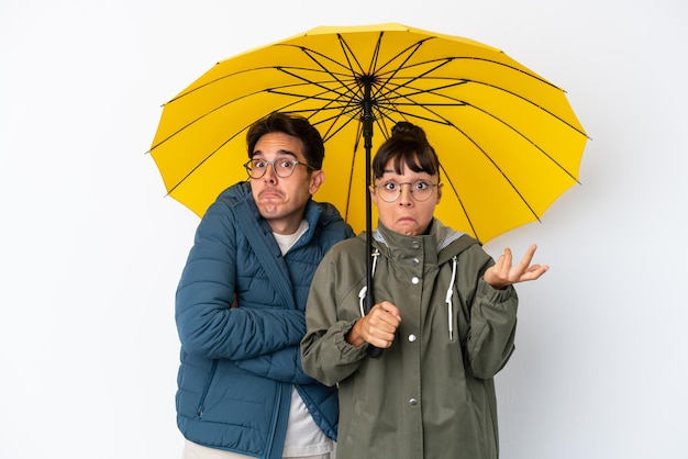 Joven pareja de raza mixta sosteniendo un paraguas aislado de fondo blanco haciendo gestos de duda mientras levanta los hombros