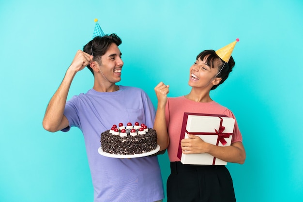 Joven pareja de raza mixta sosteniendo pastel de cumpleaños y presente aislado en fondo azul celebrando una victoria en posición de ganador