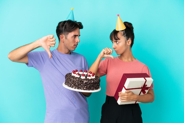 Joven pareja de raza mixta sosteniendo pastel de cumpleaños y presente aislado en fondo azul mostrando el pulgar hacia abajo con expresión negativa