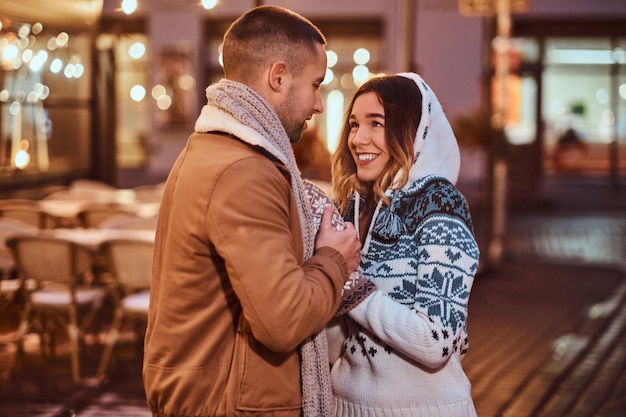 Una joven pareja romántica con ropa abrigada tomándose de la mano y calentándose mientras está de pie en la calle de la noche en Navidad