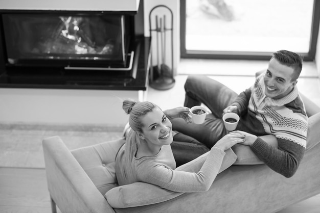 Joven pareja romántica sentada en un sofá frente a la chimenea en casa, mirándose, hablando y bebiendo café el día de otoño