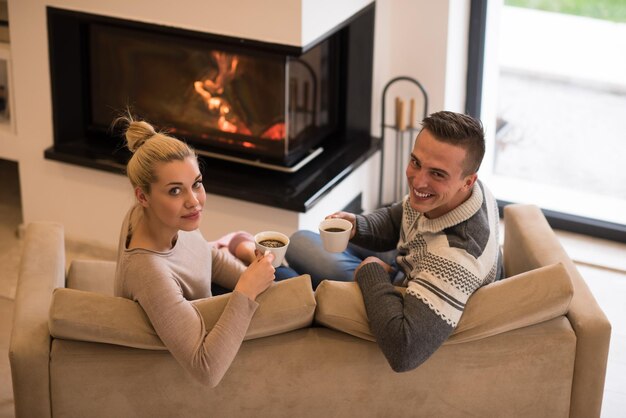 Joven pareja romántica sentada en un sofá frente a la chimenea en casa, mirándose, hablando y tomando café en el día de otoño