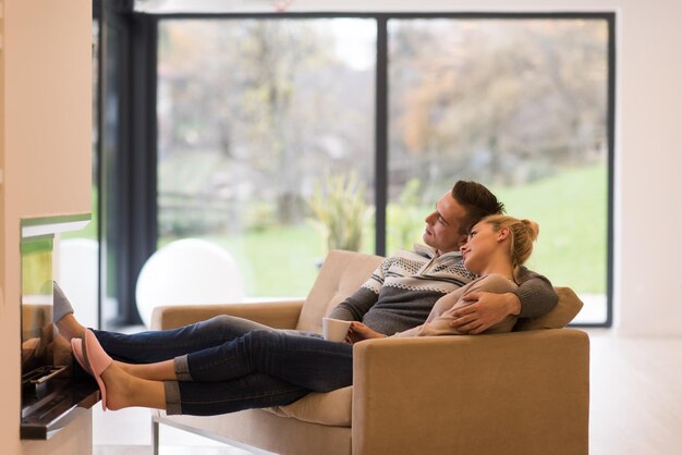 Joven pareja romántica sentada en un sofá frente a la chimenea en casa, mirándose, hablando y tomando café en el día de otoño