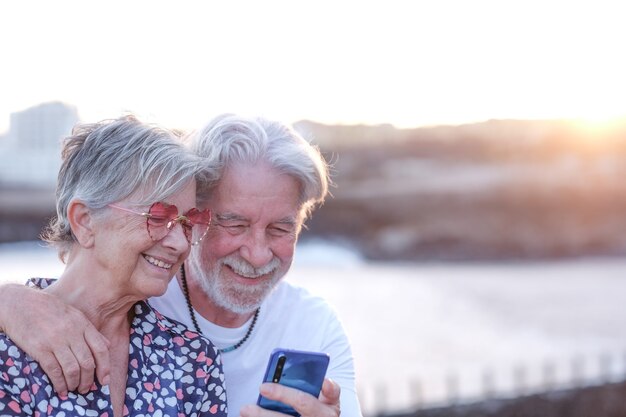 Joven pareja senior abrazando al aire libre en el mar al atardecer mediante teléfono móvil. Caucásico jubilado disfrutando de relax y estilo de vida feliz