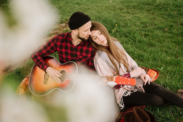 Joven pareja sentada bajo el gran árbol y el hombre tocando la guitarra