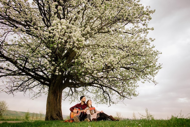 Joven pareja sentada bajo el gran árbol y el hombre tocando la guitarra