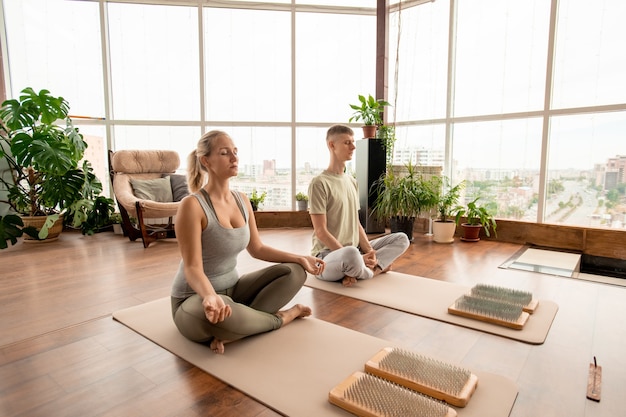 Joven pareja serena en ropa deportiva sentada en pose de loto sobre colchonetas mientras practica ejercicios de meditación juntos en el entorno hogareño