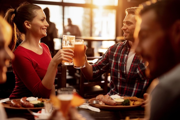 Joven pareja sonriente celebrando y brindando con cerveza mientras almuerza con amigos en un pub