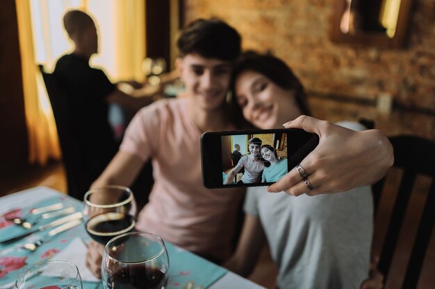 Una joven pareja sonriente sentada en un restaurante, bebiendo vino - Una joven pareja feliz tomando un selfie con un teléfono inteligente.