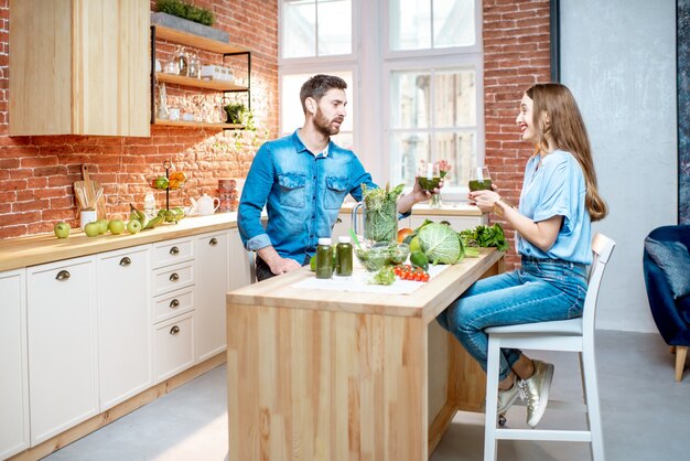 Joven pareja de vegetarianos bebiendo batido en la mesa llena de productos verdes frescos en la cocina de casa