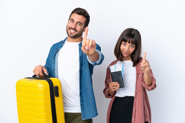 Joven pareja de viajeros sosteniendo una maleta y un pasaporte aislado de fondo blanco mostrando y levantando un dedo