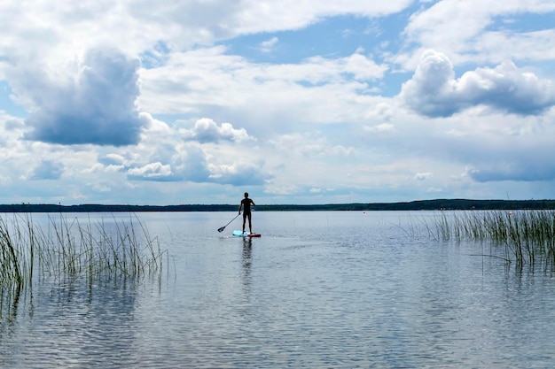 Joven de la parte de atrás en traje térmico negro paddleboarding en azul stand up paddle board en el lago contra el fondo del cielo nublado