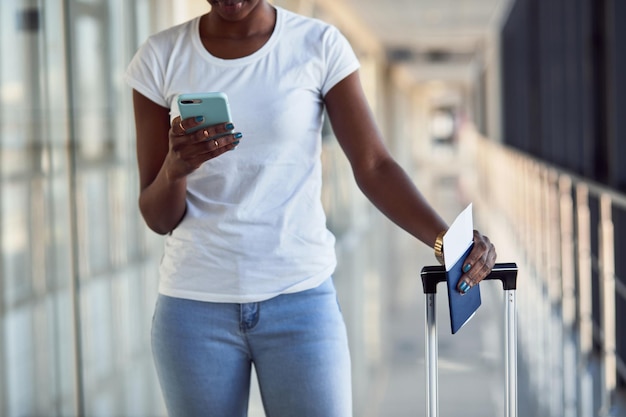 Una joven pasajera afroamericana con ropa informal está en el aeropuerto con boletos y teléfono.