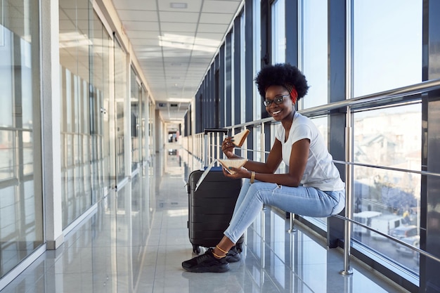 Una joven pasajera afroamericana con ropa informal está en el aeropuerto con equipaje comiendo algo.