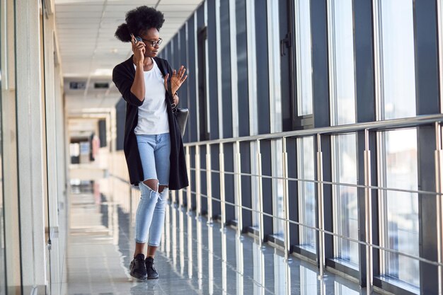 Una joven pasajera afroamericana con ropa informal está en el aeropuerto escuchando música con auriculares.