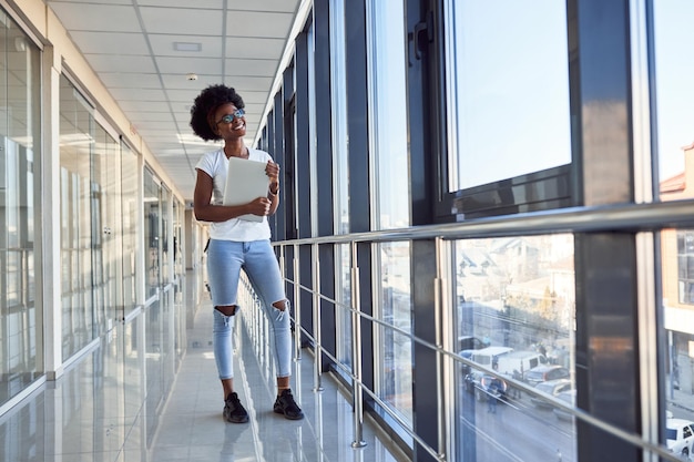 Una joven pasajera afroamericana con ropa informal está en el aeropuerto con una laptop.